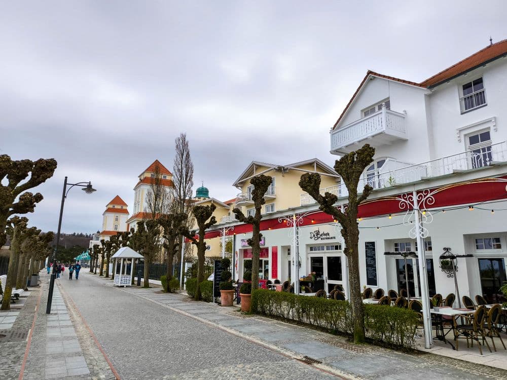 Eine kopfsteingepflasterte Straße in Binz, gesäumt von beschnittenen Bäumen und Straßencafés, wird von weißen Gebäuden mit roten Dächern unter einem bewölkten Himmel gesäumt. In der Ferne spazieren Menschen und schaffen eine entspannte Atmosphäre.