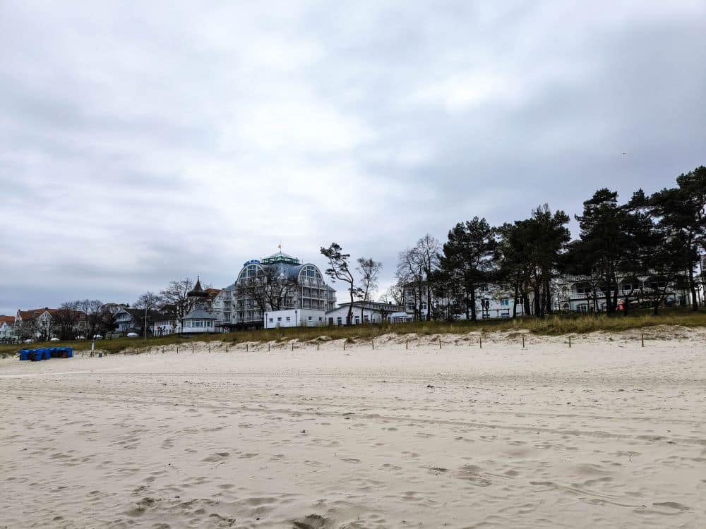 Ein Sandstrand unter bewölktem Himmel in Binz, mit mehreren großen, weißen Gebäuden und Bäumen im Hintergrund; einige Strandkörbe sind auf der linken Seite gruppiert.