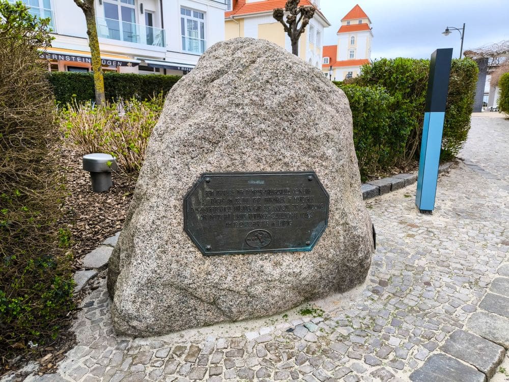 Ein großer Stein mit einer Metalltafel steht auf einem Kopfsteinpflasterweg auf dem Dorfplatz von Binz, umgeben von gestutzten Büschen und modernen Gebäuden im Hintergrund.