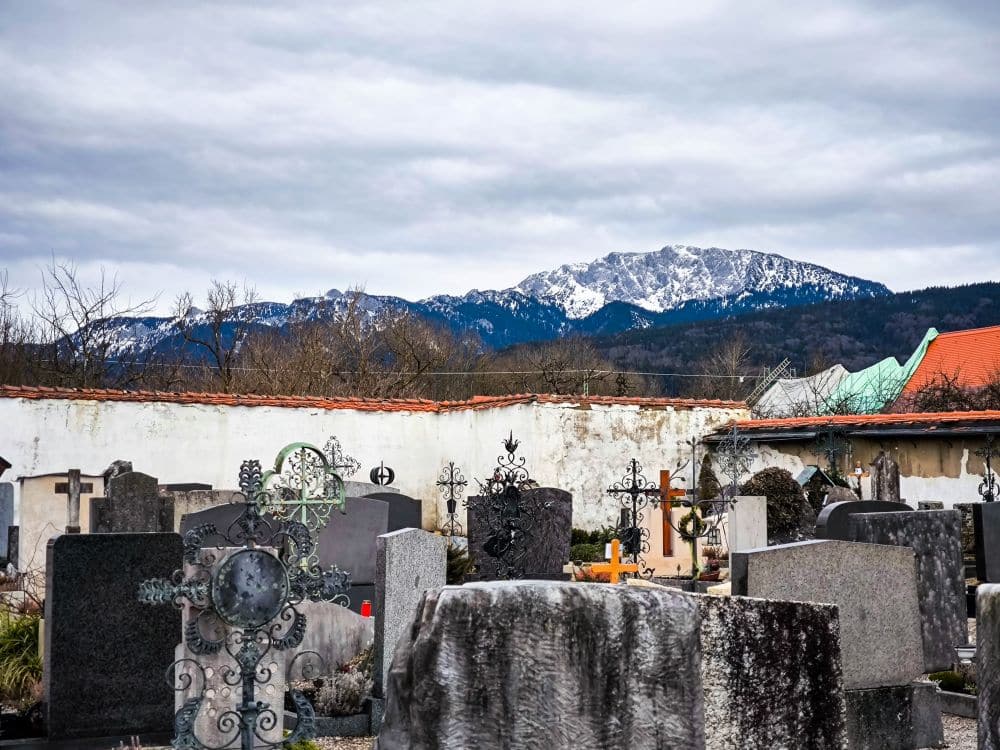 Ein Friedhof mit verschiedenen Grabsteinen und Kreuzen im Vordergrund, vor schneebedeckten Bergen und wolkenverhangenem Himmel. In der Nähe befinden sich eine Steinmauer und Barockgebäude, die möglicherweise zum Kloster Benediktbeuern gehören, mit roten und grünen Dächern.