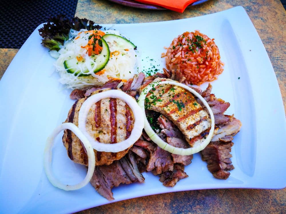 Ein Teller aus einem der Erfurter Lieblingsrestaurants: gegrillte Fleischscheiben, eine Fleischfrikadelle mit Zwiebelringen, Krautsalat mit Gurke und Tomatenreis mit Kräutern, alles auf einem weißen, quadratischen Teller.
