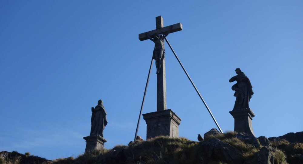 Drei Statuen stehen auf einem grasbewachsenen Hügel vor einem strahlend blauen Himmel: in der Mitte ein Kruzifix mit Jesus, flankiert von zwei Figuren auf Sockeln. Diese heitere Szene erwartet Wanderer, die die Region Wandern Rhön bei Milseburg erkunden.
