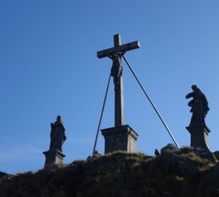 Drei Statuen stehen auf einem grasbewachsenen Hügel vor einem strahlend blauen Himmel: in der Mitte ein Kruzifix mit Jesus, flankiert von zwei Figuren auf Sockeln. Diese heitere Szene erwartet Wanderer, die die Region Wandern Rhön bei Milseburg erkunden.