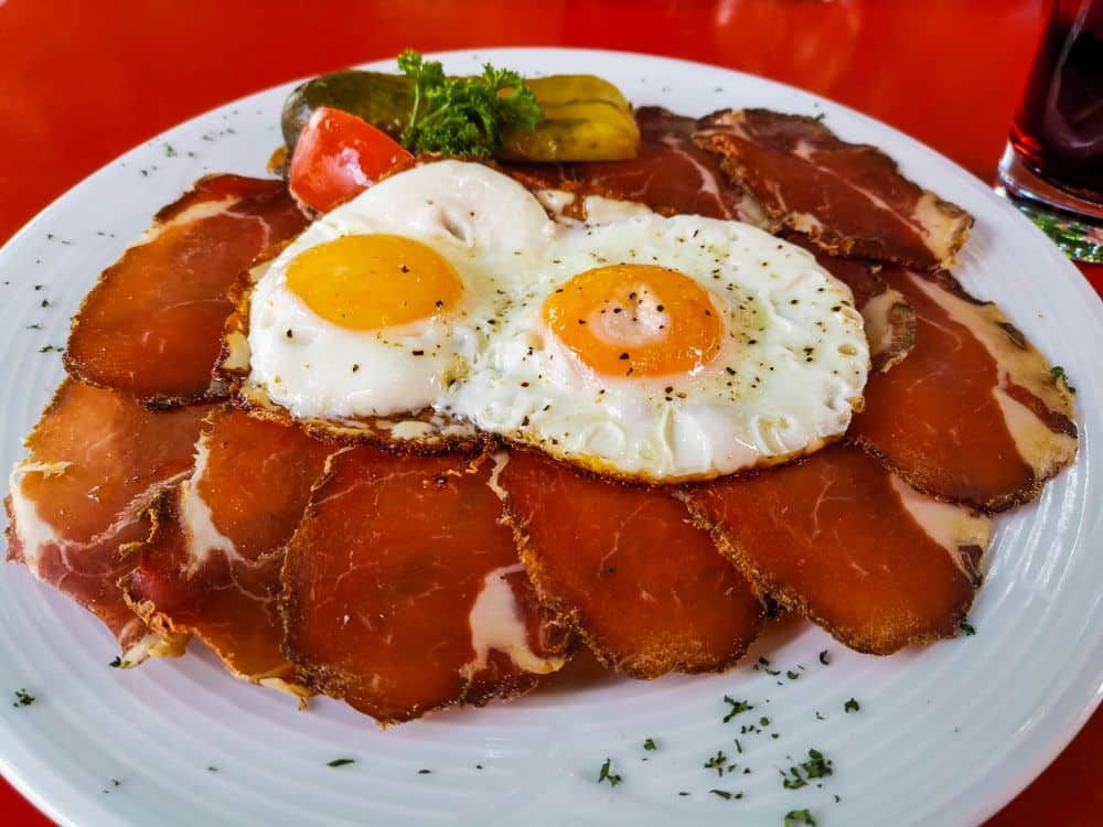 Ein weißer Teller mit zwei Spiegeleiern auf dünnen Pökelfleischscheiben, garniert mit Kräutern, einer Essiggurke, Petersilie und einer Tomatenscheibe, steht auf einem roten Tisch - typisch für manche Lieblingsrestaurants in Erfurt.