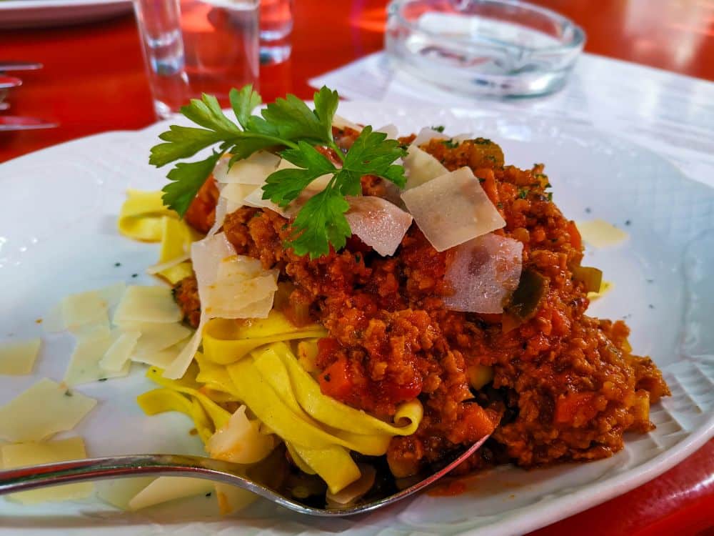 Ein Teller mit Tagliatelle-Nudeln, die mit einer reichhaltigen Fleisch-Tomatensoße übergossen und mit frischer Petersilie und gehobeltem Parmesan garniert sind, erinnert an den Geschmack von Erfurt. Ein Löffel ruht auf dem Tellerrand.