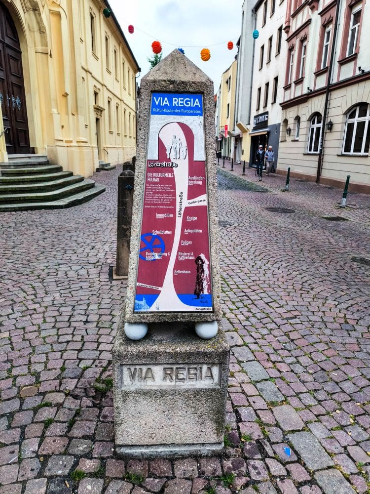 Ein Stein mit der Aufschrift "VIA REGIA" steht auf einer Kopfsteinpflasterstraße in Fulda. Darüber befindet sich eine bunte Informationstafel über die historische europäische Route. Im Hintergrund säumen Gebäude die Straße.