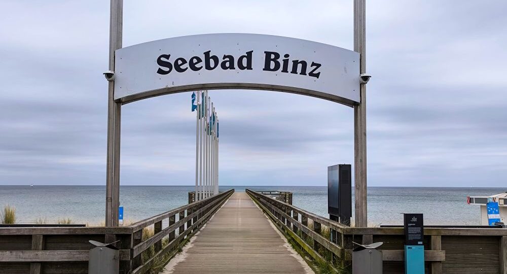 Eine hölzerne Seebrücke in Binz führt zum Meer, eingerahmt von einem großen Schild "Seebad Binz". Fahnen säumen den Steg, und der bedeckte Himmel schafft eine ruhige und heitere Küstenszene.