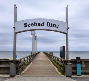 Eine hölzerne Seebrücke in Binz führt zum Meer, eingerahmt von einem großen Schild "Seebad Binz". Fahnen säumen den Steg, und der bedeckte Himmel schafft eine ruhige und heitere Küstenszene.