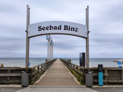 Eine hölzerne Seebrücke in Binz führt zum Meer, eingerahmt von einem großen Schild "Seebad Binz". Fahnen säumen den Steg, und der bedeckte Himmel schafft eine ruhige und heitere Küstenszene.