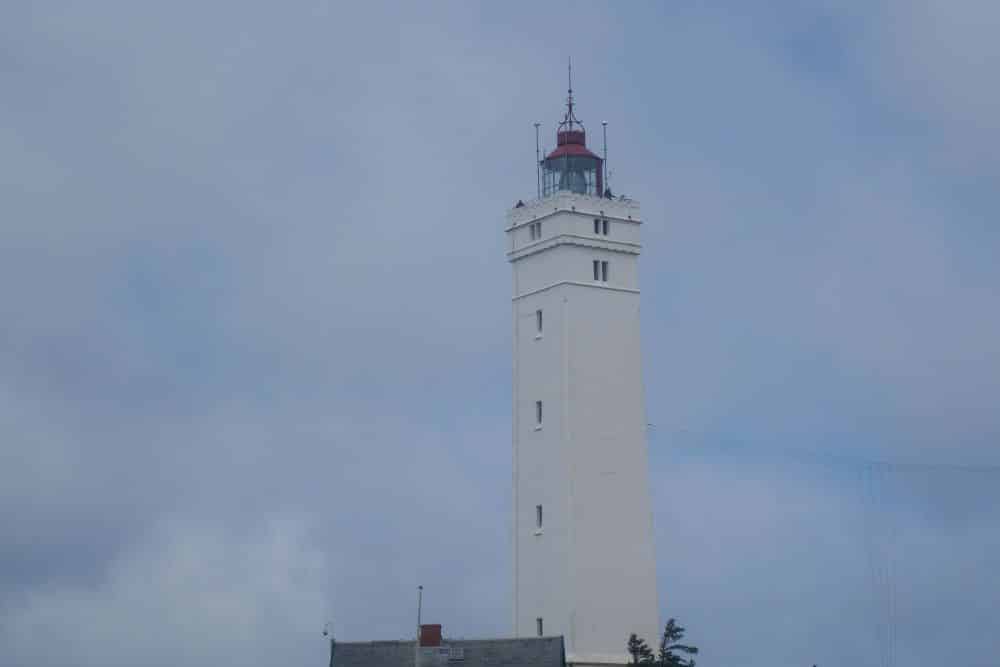 Ein hoher, weißer Leuchtturm mit roter Spitze erhebt sich vor einem wolkenverhangenen blauen Himmel in Blavand, Dänemark. Antennen und Fenster zieren seine Struktur, während ein Teil eines grauen Daches ihn im wildromantischen Westen verankert.