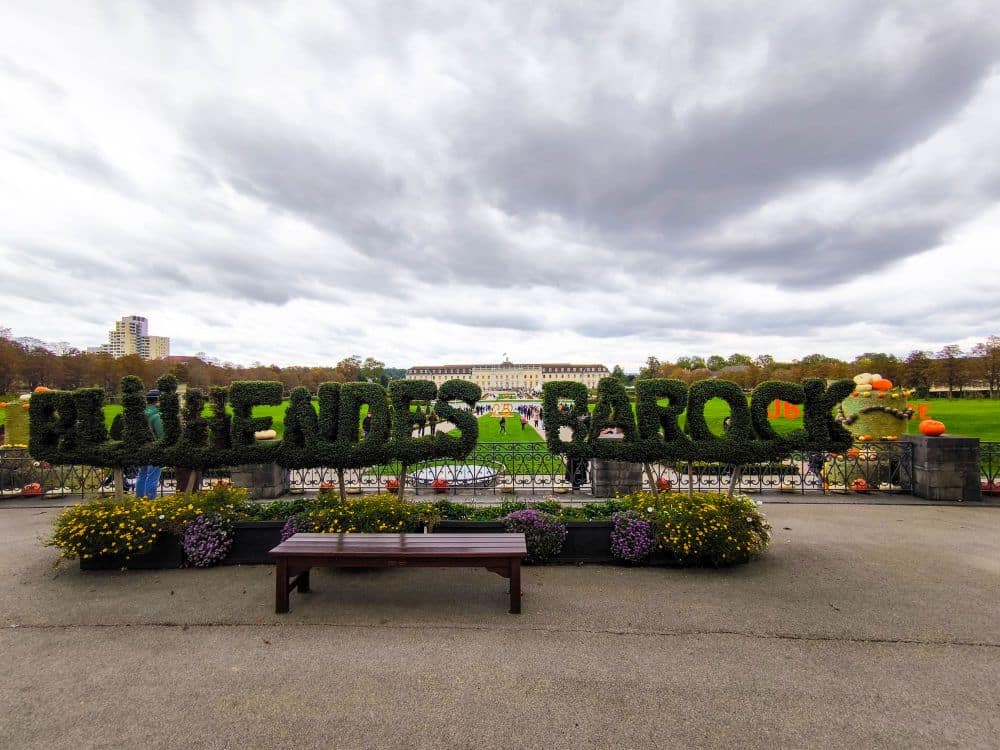 Ein Park in Ludwigsburg mit Gärten aus gepflegten Hecken mit dem Schriftzug BLÜHENDES BAROCK, bunten Blumenbeeten, einer Holzbank davor und einem großen historischen Gebäude, das unter einem bewölkten Himmel die Barocke Pracht zeigt.