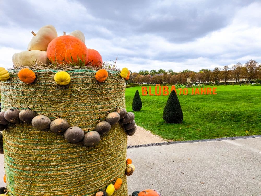 Ein großer, mit Kürbissen und Kürbissen geschmückter Heuballen steht in einem Park. Im Hintergrund steht in roten Buchstaben BLUBA 70 JAHRE auf der Wiese in den Barockgärten von Ludwigsburg, mit gestutzten Büschen und wolkenverhangenem Himmel über uns.