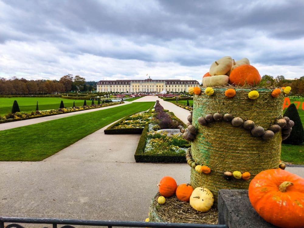 Ein gepflegter Garten mit ordentlich geschnittenen Rasenflächen und Blumenbeeten führt zu einem großen, eleganten Ludwigsburger Gebäude. Im Vordergrund erinnern mit Kürbissen und Kürbissen geschmückte Heuballen an die barocke Pracht unter einem wolkenverhangenen Himmel.