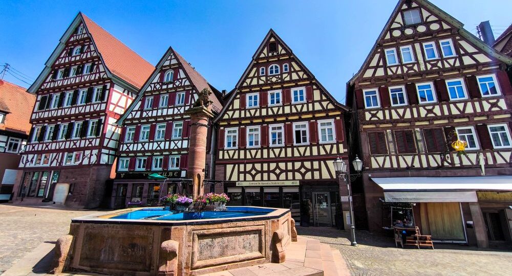 Bunte Fachwerkhäuser mit steilen Dächern und dekorativen Fassaden säumen einen kopfsteingepflasterten Platz in Calw, in dessen Mitte ein steinerner Brunnen mit einem Blumenarrangement steht, unter einem klaren blauen Himmel.
