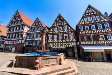 Bunte Fachwerkhäuser mit steilen Dächern und dekorativen Fassaden säumen einen kopfsteingepflasterten Platz in Calw, in dessen Mitte ein steinerner Brunnen mit einem Blumenarrangement steht, unter einem klaren blauen Himmel.