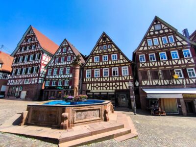 Bunte Fachwerkhäuser mit steilen Dächern und dekorativen Fassaden säumen einen kopfsteingepflasterten Platz in Calw, in dessen Mitte ein steinerner Brunnen mit einem Blumenarrangement steht, unter einem klaren blauen Himmel.