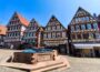 Bunte Fachwerkhäuser mit steilen Dächern und dekorativen Fassaden säumen einen kopfsteingepflasterten Platz in Calw, in dessen Mitte ein steinerner Brunnen mit einem Blumenarrangement steht, unter einem klaren blauen Himmel.