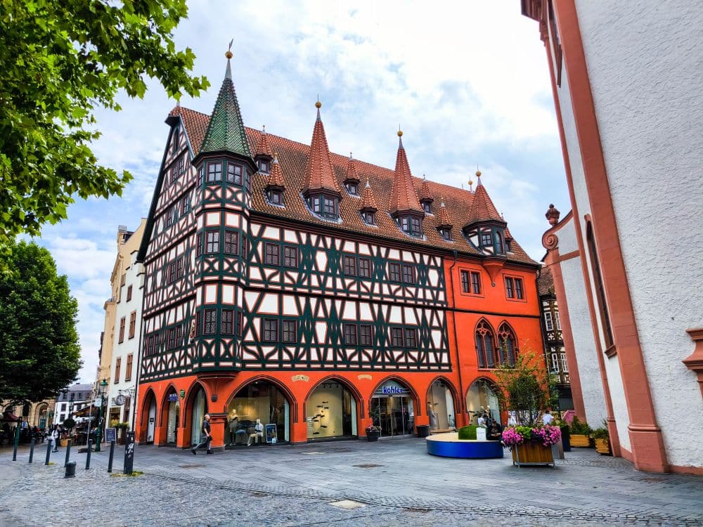 Ein traditionelles Fachwerkhaus mit roten Akzenten und spitzen Dächern steht auf einem kopfsteingepflasterten Platz in Fulda, umgeben von Bäumen, Blumenkübeln und Geschäften auf Straßenebene unter einem teilweise bewölkten Himmel.