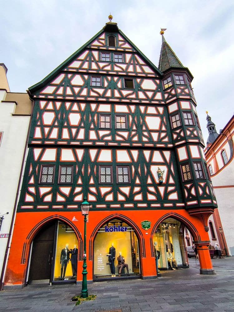 Ein auffälliges, verziertes Fachwerkgebäude in Fulda mit schwarz-rot gemustertem Holzwerk und gewölbten Schaufenstern auf Straßenebene. Die oberen Stockwerke sind mit einem komplizierten Kreuzmuster versehen. Im Erdgeschoss ist ein Bekleidungsgeschäft zu sehen.