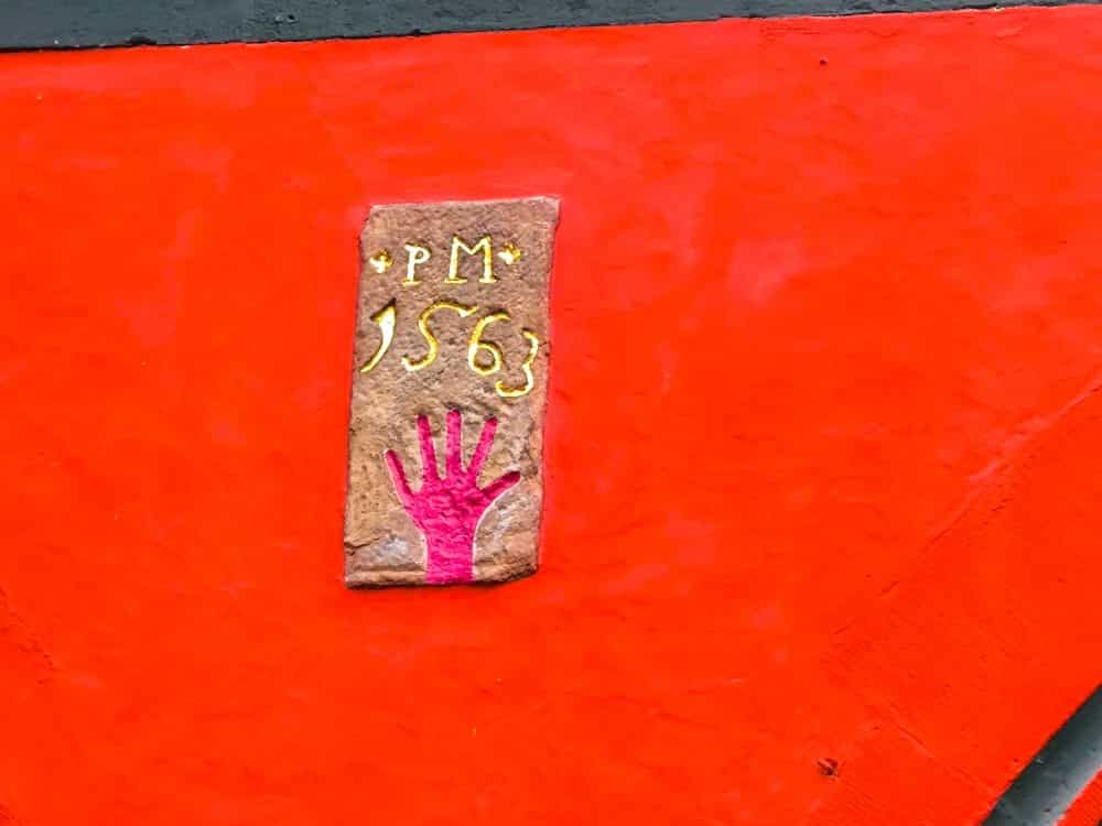 An einer roten Wand in Fulda befindet sich eine kleine rechteckige Plakette mit der gelben Aufschrift "PM 1563" und einem rosafarbenen Handabdruck unter dem Text. Die Tafel hebt sich von dem leuchtend orange-roten Hintergrund ab.