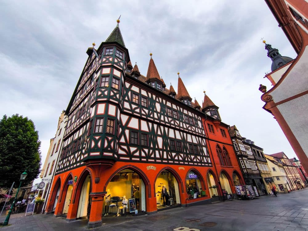 Ein hohes, verziertes Fachwerkgebäude mit roten Bögen und mehreren spitzen Türmen erhebt sich unter einem wolkenverhangenen Himmel in Fulda, an einer belebten Straße mit Geschäften und Fußgängern.