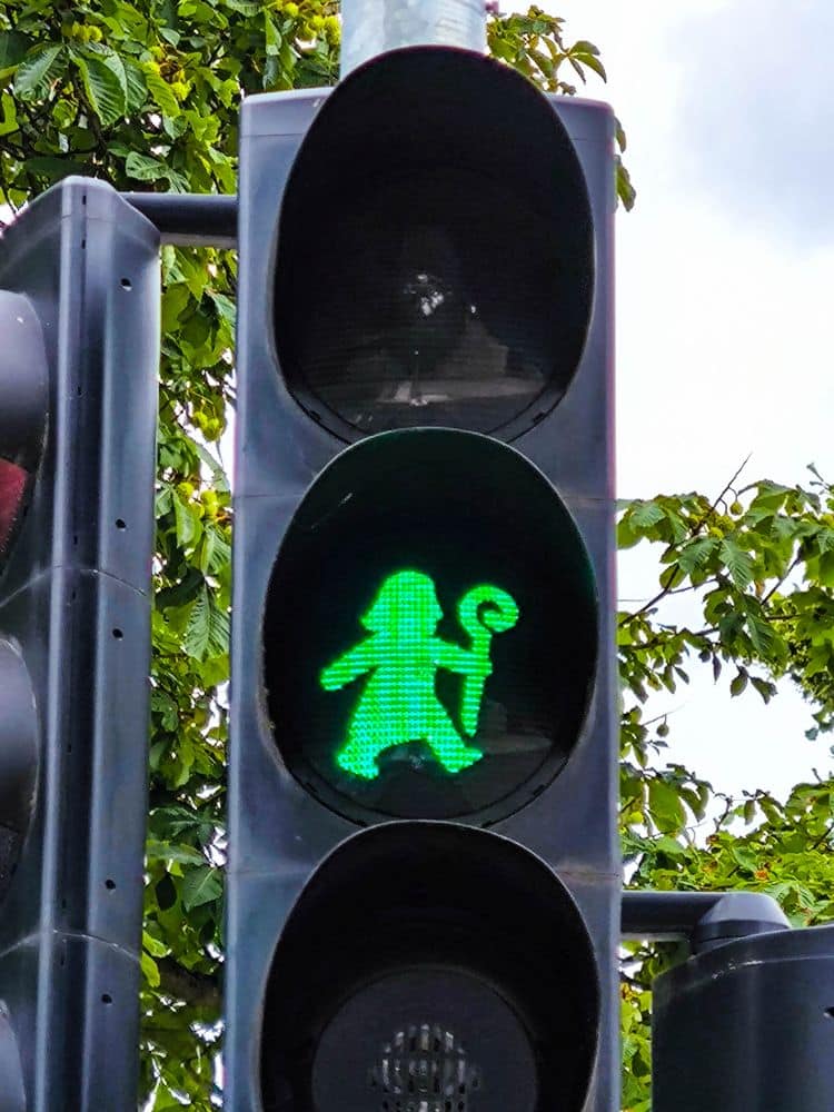 Eine grüne Ampel in Fulda zeigt eine schreitende Figur, die einen Stab hält und einem Zauberer ähnelt, vor einem bewaldeten Hintergrund und einem bewölkten Himmel.