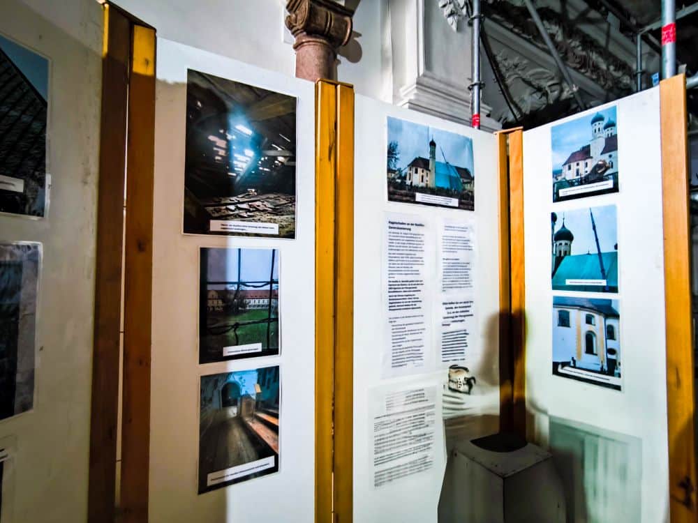 Eine Schautafel mit Fotos von Barockgebäuden und architektonischen Stätten, darunter auch das Kloster Benediktbeuern, steht im Innenbereich unter heller Beleuchtung. Holztafeln rahmen die Ausstellung ein, im Hintergrund sind eine Säule und ein Blick auf Berge zu sehen.