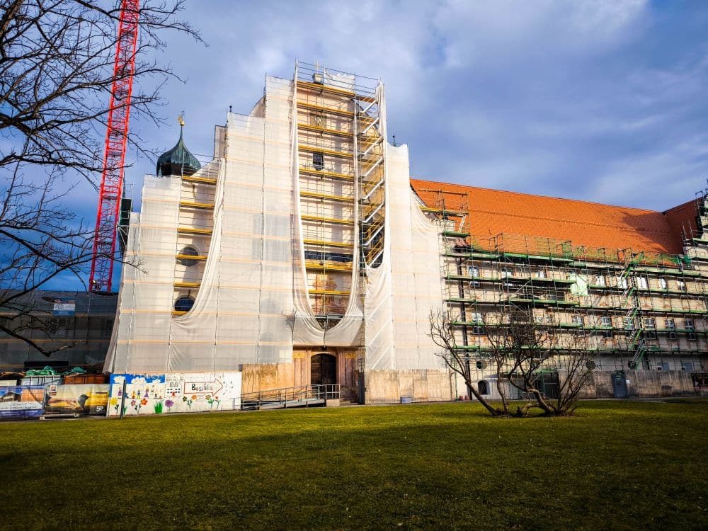 Ein historisches Barockgebäude im Kloster Benediktbeuern, das mit Gerüsten und weißen Planen verkleidet ist, wird gerade renoviert. Auf der linken Seite steht ein roter Kran. Davor stehen ein Baum und eine Wiese, und die Berge erheben sich unter einem teilweise bewölkten Himmel.