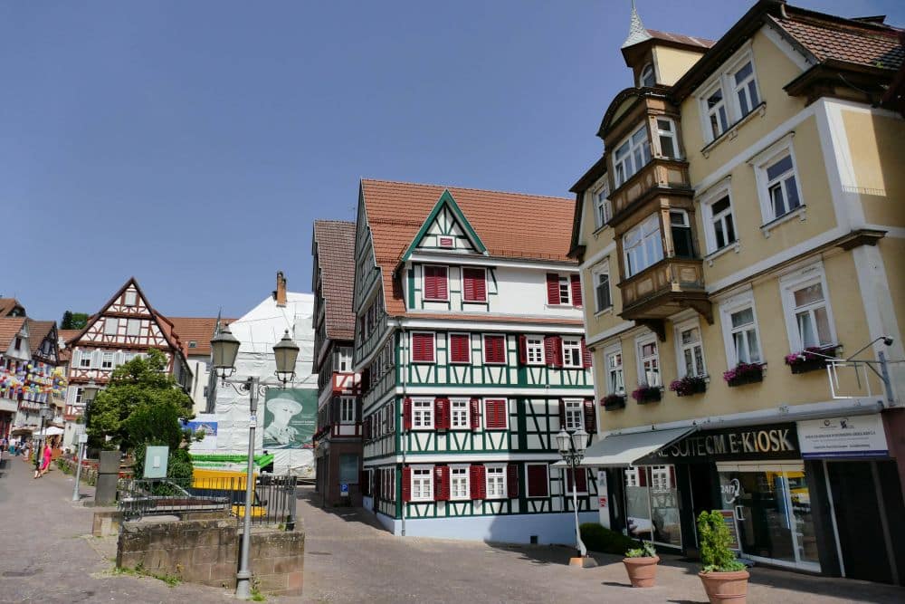 Eine sonnige Straße in Calw mit bunten Fachwerkhäusern, Blumenkästen an den Fenstern und einem kleinen Kiosk. Die Fußgänger schlendern bei strahlend blauem Himmel über den Kopfsteinpflasterweg.