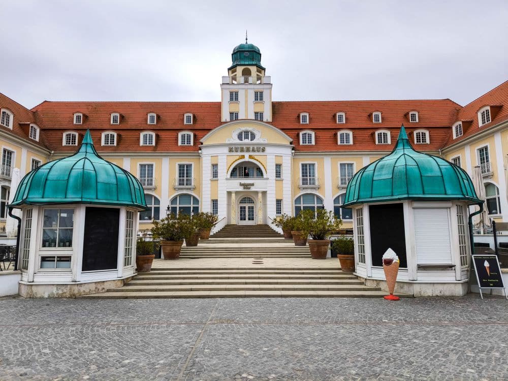 Ein großes, elegantes Gebäude in Binz mit rotem Dach und einem zentralen Uhrenturm, flankiert von zwei Kiosken mit grünen Kuppeln. Eine breite Treppe führt zum Eingang, vor dem sich Topfpflanzen und eine Eistütenstatue befinden.