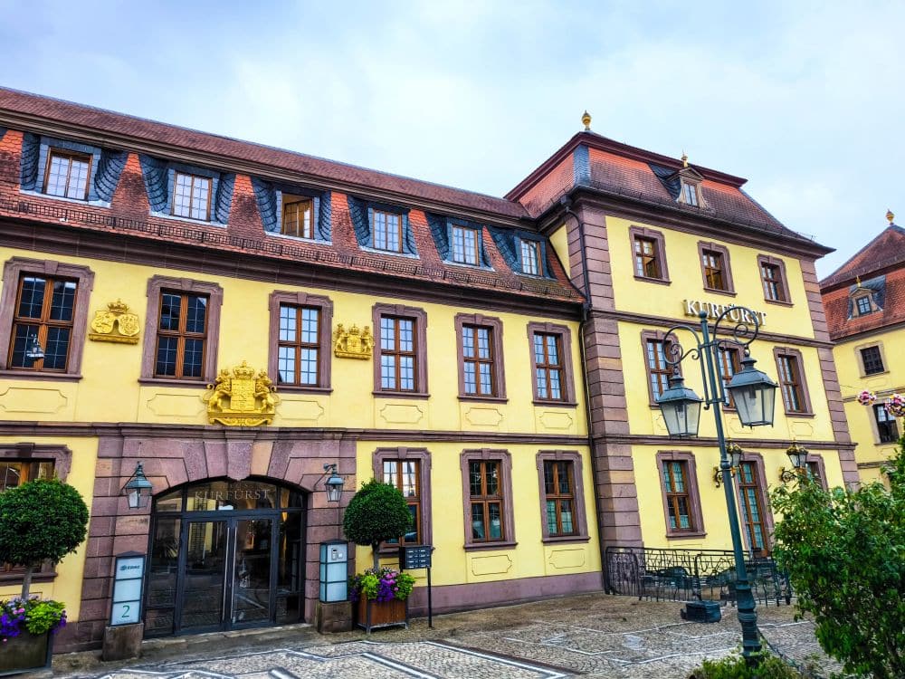 Ein prächtiges gelbes und rosafarbenes historisches Gebäude in Fulda mit vielen Fenstern, dekorativen goldenen Wappen und Schildern mit der Aufschrift KURFÜRST. Verschnörkelte Straßenlaternen und Grünpflanzen schmücken den gepflasterten Innenhof davor.