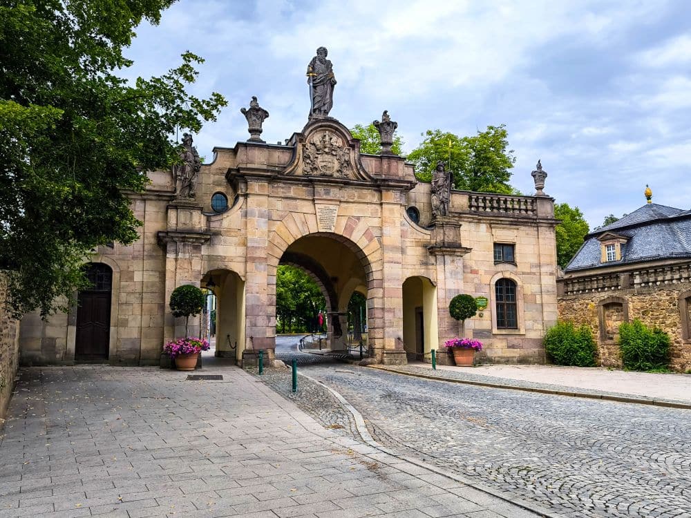 Ein historischer steinerner Torbogen in Fulda, geschmückt mit Statuen und dekorativen Schnitzereien, steht über einer Straße mit Kopfsteinpflaster, flankiert von Topfpflanzen und Bäumen, unter einem bewölkten Himmel. Auf der rechten Seite ist ein kleines Steingebäude zu sehen.