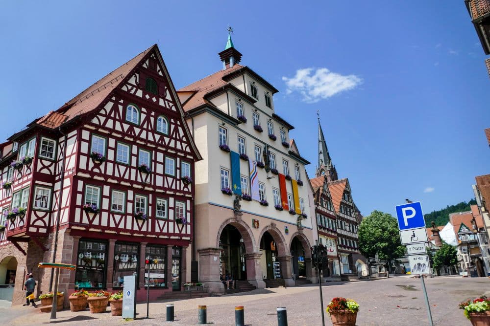 Ein malerischer europäischer Stadtplatz in Calw mit farbenfrohen Fachwerkhäusern, einem geschmückten Rathaus mit Blumenkästen und einem hohen Kirchturm, der sich im Hintergrund unter einem klaren blauen Himmel erhebt.