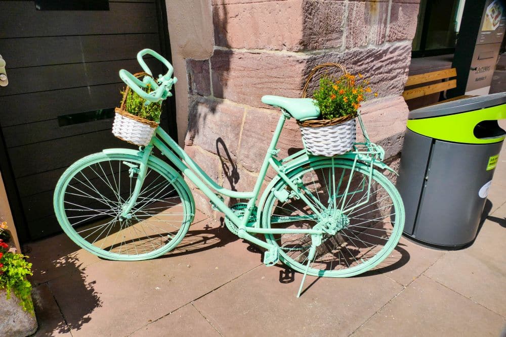 Ein mintgrünes Fahrrad mit weißen Körben, die orangefarbene Blumen tragen, ist an einem sonnigen Tag an einer Steinmauer in Calw geparkt.