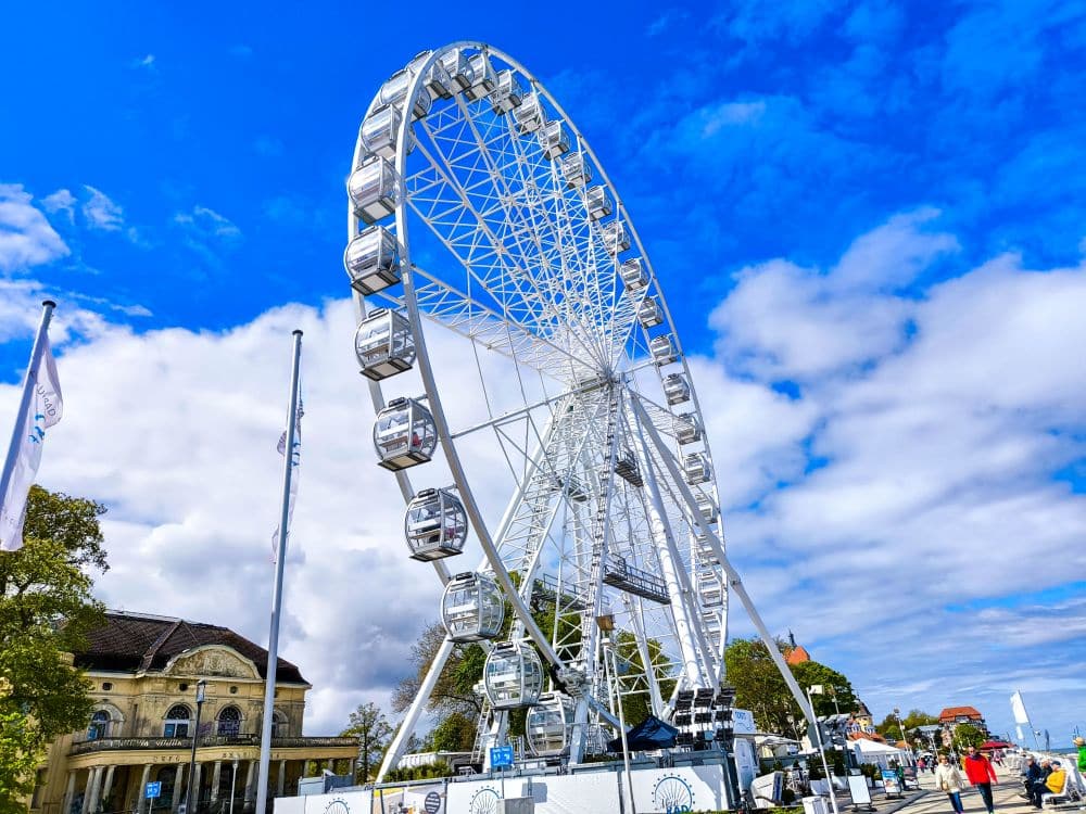 Ein großes weißes Riesenrad steht vor einem strahlend blauen Himmel mit vereinzelten Wolken in Kühlungsborn, umgeben von Menschen, Gebäuden und Bäumen an einem sonnigen Ostseetag - perfekt für eine Auszeit.