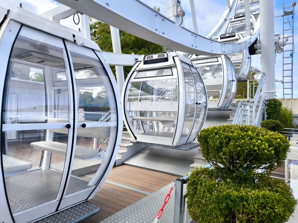 Eine Nahaufnahme mehrerer moderner, weißer Riesenradkabinen mit klaren Fenstern in der Nähe einer Einstiegsplattform, umgeben von grünen Büschen und Bäumen - perfekt für eine Auszeit in Kühlungsborn an der Ostsee.