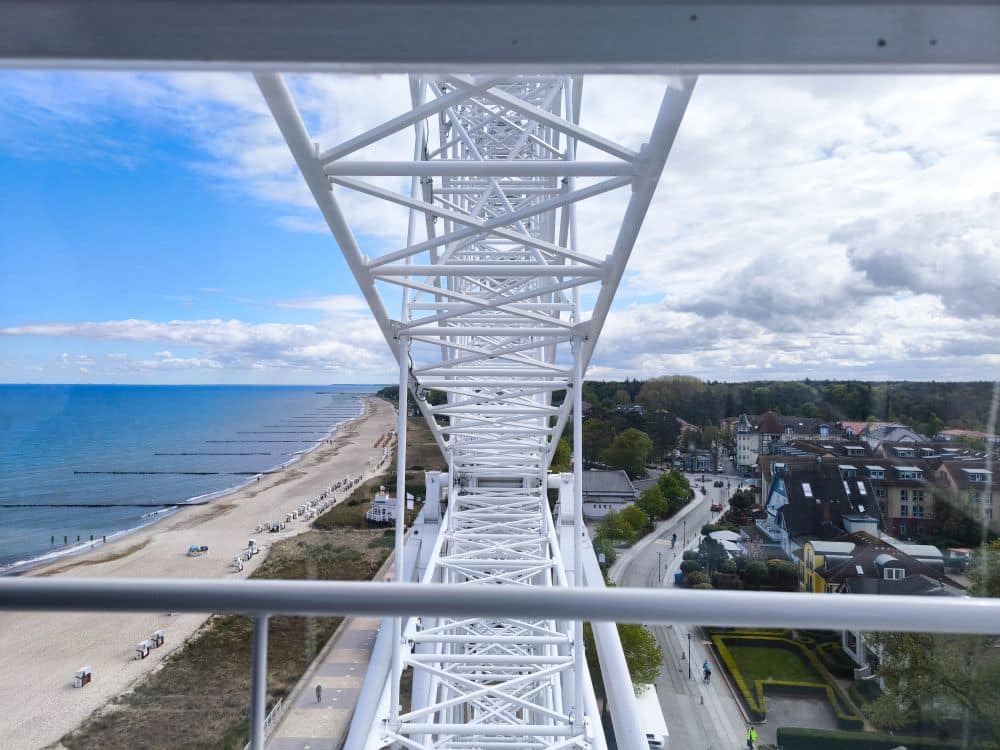 Blick aus dem Inneren eines Riesenrads, das seine weiße Metallstruktur zeigt. Unten erstreckt sich Kühlungsborns Sandstrand an der Ostsee, mit Spaziergängern und einer Stadt am Meer unter einem teilweise bewölkten Himmel - perfekt für eine Auszeit.