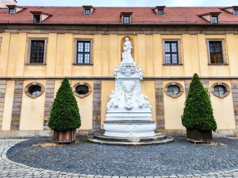Eine weiße, verzierte Statue mit mehreren Figuren steht zwischen zwei hohen, kegelförmig geschnittenen Büschen vor einem gelben Fulda-Gebäude mit runden und rechteckigen Fenstern und einem roten Ziegeldach. Kopfsteinpflaster umgibt die Statue.