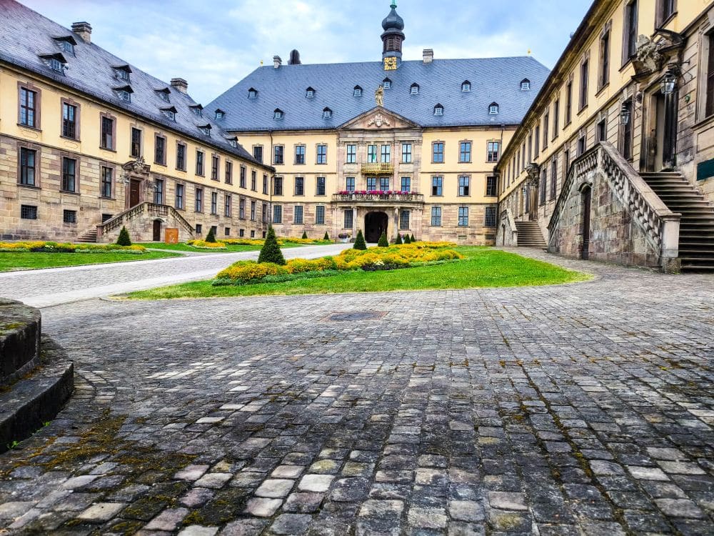 Ein großes, historisches Gebäude in Fulda mit einem gepflasterten Innenhof, gepflegten Gärten und symmetrischen Steintreppen auf beiden Seiten. Das Gebäude verfügt über zahlreiche Fenster und einen zentralen Teil, der von einem Uhrenturm gekrönt wird.
