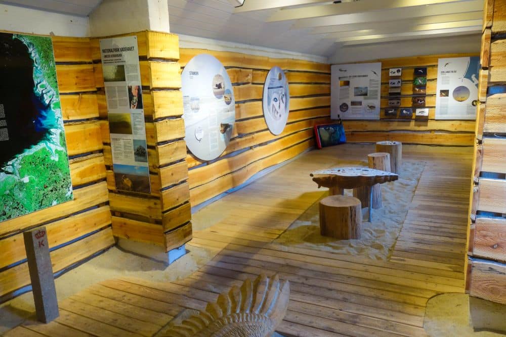 Ein hölzerner Ausstellungsraum mit sandigem Bodenbelag erinnert an den wildromantischen Charme von Dänemarks Westen. Informationstafeln und rustikale Bänke bieten einen einladenden, hellen Raum, der vom Küstengeist Blavands inspiriert ist.