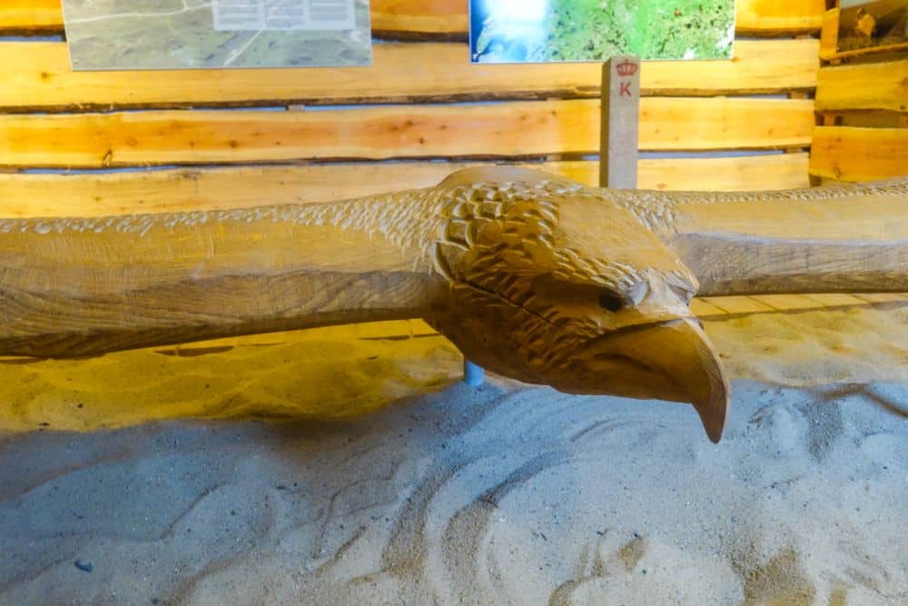 Eine große Holzskulptur eines Adlers mit ausgebreiteten Flügeln steht auf einem Sandboden in einem rustikalen Raum mit Holzwänden und Informationstafeln, die an den wildromantischen Geist von Dänemarks Westen bei Blavand erinnern.