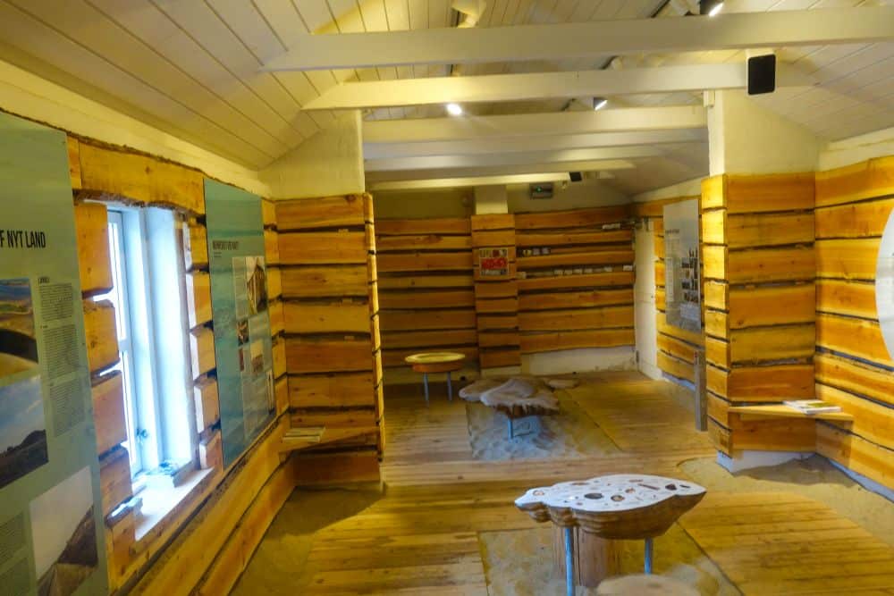 Ein überdachter Ausstellungsraum in Blavand mit Holzwänden und -böden, Informationstafeln, natürlichem Licht aus einem Fenster und interaktiven Tischen schafft eine wildromantische und lehrreiche Atmosphäre, die von Dänemarks Westen inspiriert ist.