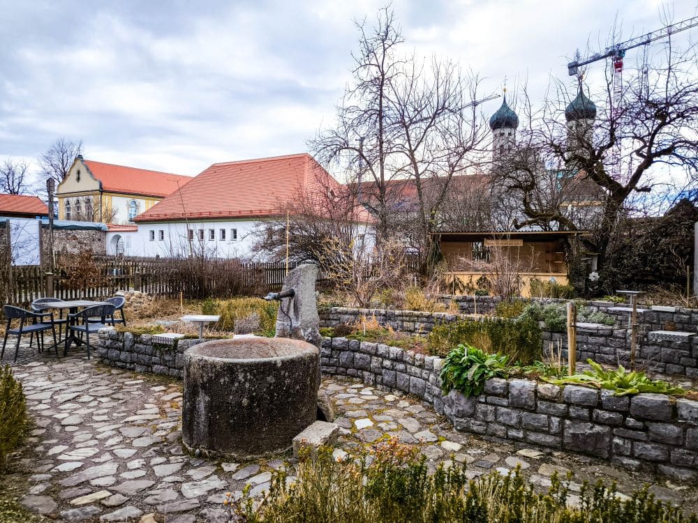 Ein kleiner Steinhof mit einem Brunnen und Tischen im Freien, umgeben von blattlosen Bäumen und Gartenbeeten. Im Hintergrund erheben sich die Barocktürme des Klosters Benediktbeuern und rotgedeckte Gebäude unter einem wolkenverhangenen Himmel gegen die fernen Berge.