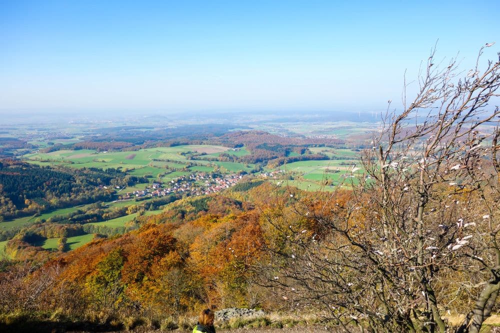 Der Blick von der Milseburg in der Rhön zeigt herbstlich gefärbte Bäume im Vordergrund, Felder und ein kleines Dorf in einem grünen Tal, darüber ein strahlend blauer Himmel - ein perfekter Ort für Ihr nächstes Wanderabenteuer.
