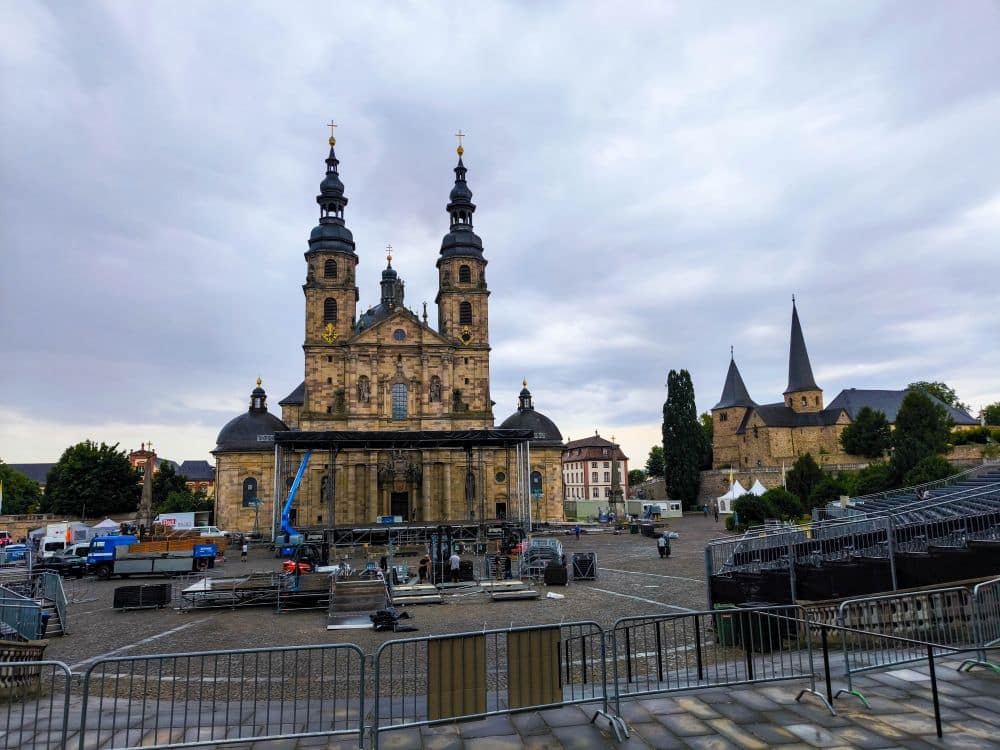 Ein großer historischer Dom mit Zwillingstürmen steht auf dem Stadtplatz von Fulda. Im Vordergrund stehen Baumaschinen und Metallbarrieren, die auf eine Veranstaltung hindeuten. Bewölkter Himmel darüber. Andere Kirchengebäude und Bäume sind in der Nähe zu sehen.