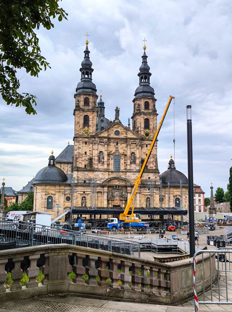 Ein großer gelber Kran arbeitet vor Fuldas historischer Barockkirche mit ihren Zwillingstürmen, inmitten von Baugeräten und Fahrzeugen auf dem Stadtplatz unter einem bewölkten Himmel.