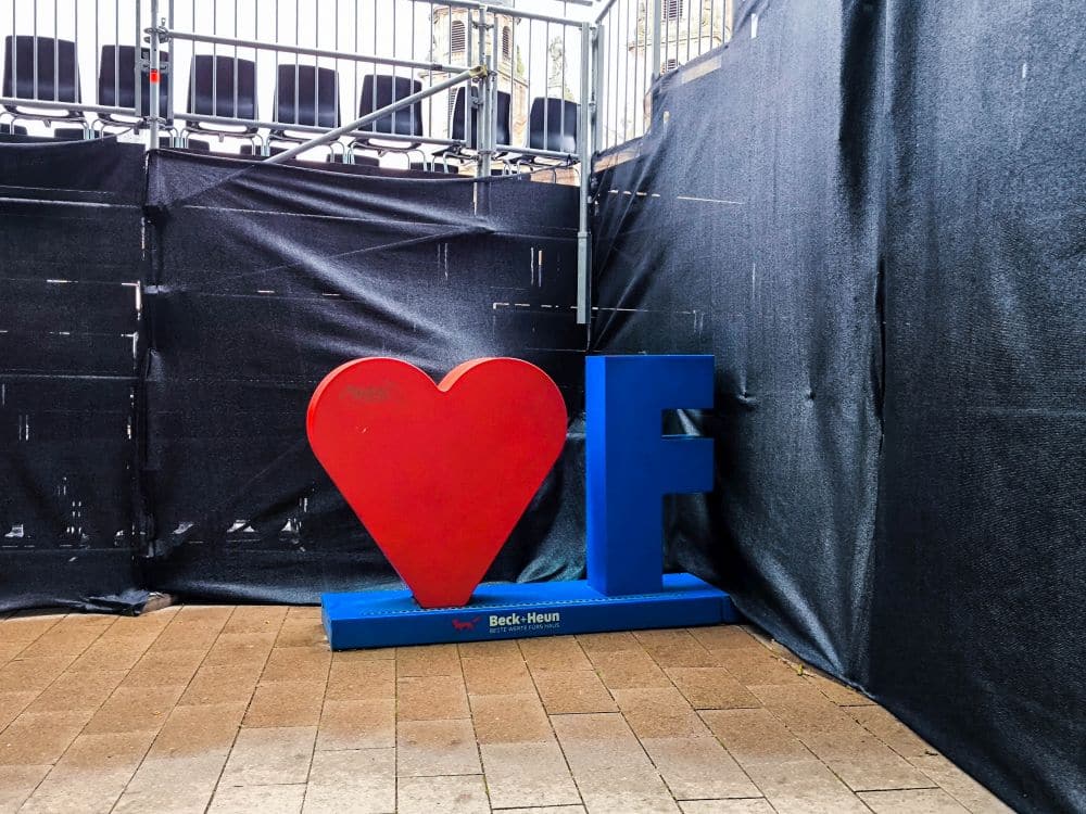 Eine große rote Herzform steht neben einem blauen Buchstaben F auf einer gepflasterten Fläche in Fulda, mit schwarzem Stoff und Metallbarrieren im Hintergrund.