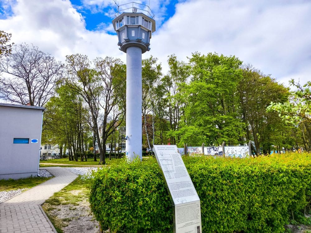 Ein hoher, weißer Wachturm steht neben einer grünen Hecke und einem Hinweisschild in Kühlungsborn, mit grünen Bäumen und einem teilweise bewölkten blauen Himmel im Hintergrund. Ein gepflasterter Weg und ein nahegelegenes Gebäude tragen zu dieser ruhigen Ostsee-Auszeit-Kulisse bei.