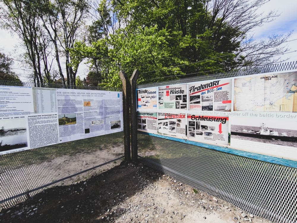 Zwei Informationstafeln mit Texten, Fotos und Karten stehen an der Ecke eines Drahtzauns inmitten von Bäumen und Grün in Kühlungsborn und laden an einem sonnigen Tag zu einer Auszeit an der Ostsee ein.