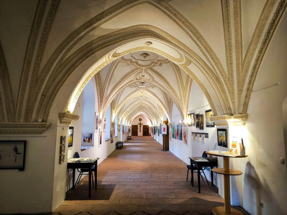 Ein langer, gewölbter Gang mit verzierten Barockgewölben, gesäumt von gerahmten Kunstwerken an beiden Wänden. An den Seiten stehen kleine Tische und Stühle, und am Ende ist ein Kreuz zu sehen - die warme Beleuchtung unterstreicht die Architektur des Klosters Benediktbeuern.
